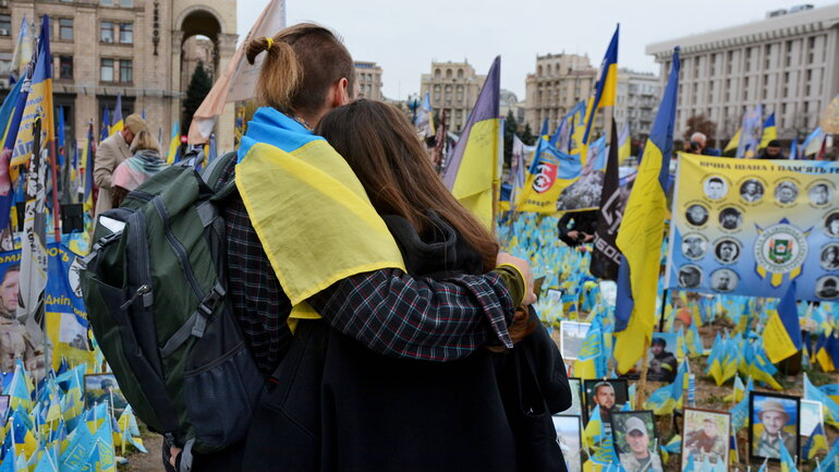 Zwei Personen umarmen sich und blicken auf eine Vielzahl ukrainischer Fahnen, die auf einem Platz aufgebaut sind.