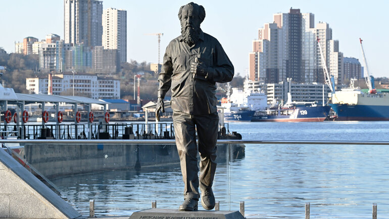 A gray statue of a man stands on the bank of a river against the background of urban apartment buildings.