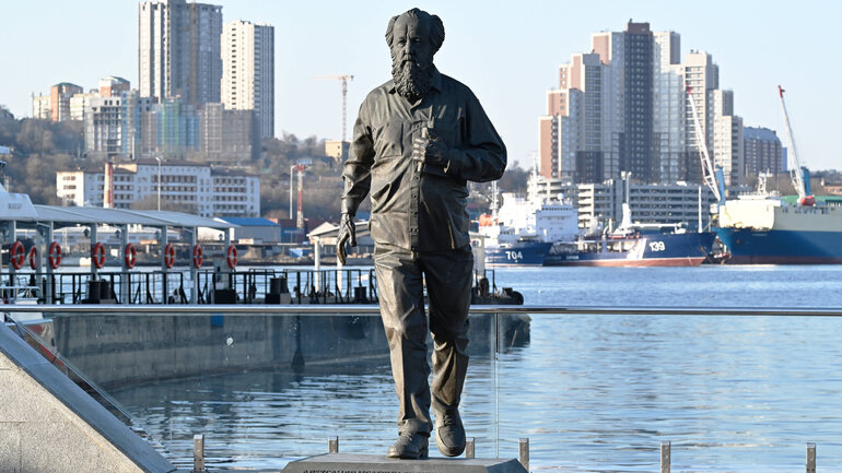 Eine graue Statue eines Mannes steht am Ufer eines Flusses vor dem Hintergrund städtischer Mehrfamilienhäuser. 