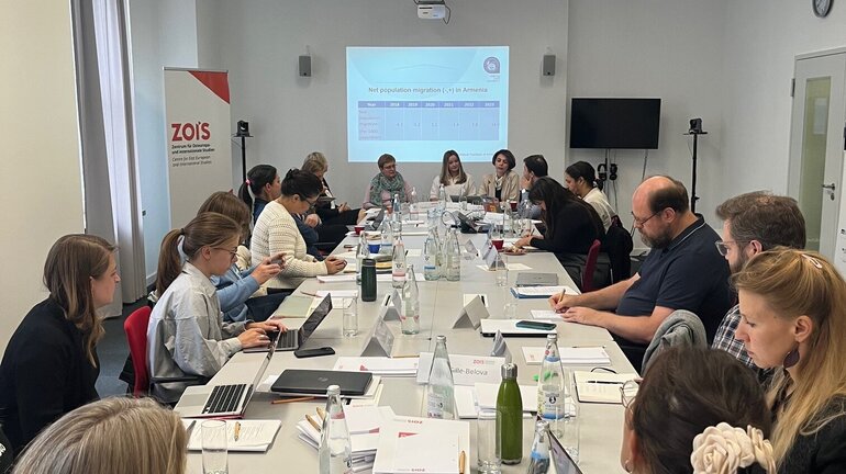 A large group of people is sitting together at a long table. Behind them, a presentation can be seen projected onto a wall.