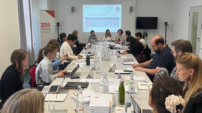 A large group of people is sitting together at a long table. Behind them, a presentation can be seen projected onto a wall.