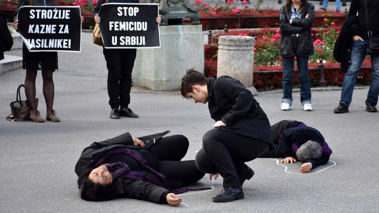 Several people dressed in black are in a square. Two of them are lying on the ground and two others in the background are holding protest placards.