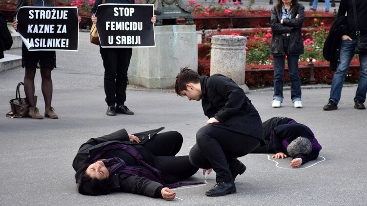 Mehrere schwarz gekleidete Personen befinden sich auf einem Platz. Zwei von ihnen liegen am Boden und zwei weitere im Hintergrund halten Protestplakate.