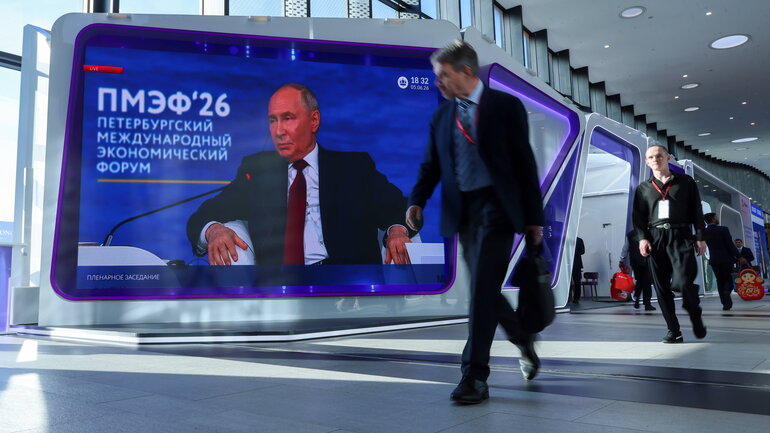 Two people walk past a large screen showing a speech by Putin at the St. Petersburg International Economic Forum.
