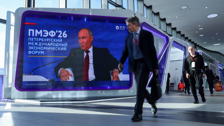 Zwei Personen laufen an einem großen Bildschirm, der eine Rede Putins auf dem internationalen Wirtschaftsforum in St. Petersburg überträgt vorbei.