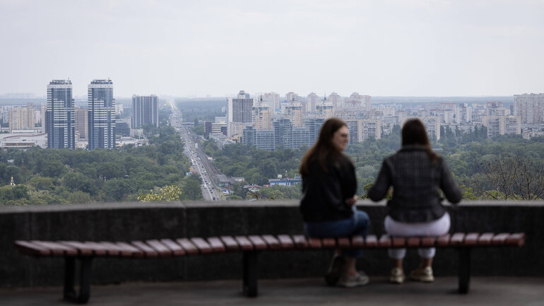 Two women are sitting on a bench facing away from the camera looking out onto a city with a few high-rise buildings.
