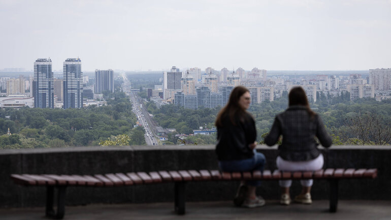 Zwei Frauen sitzen mit dem Rücken zur Kamera auf einer Bank und blicken auf eine Stadt mit einigen Hochhäusern.