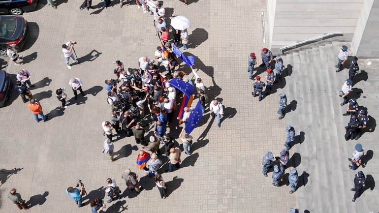 Around 20 people with Armenian and EU flags stand in front of the Armenian Foreign Ministry in Yerevan. Journalists crowd in front of them and police officers stand behind them. The Armenian and Russian foreign ministers are meeting in the foreign ministry at this time.