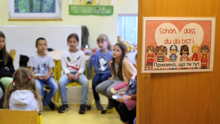 Hinter einer halbgeöffneten Tür sitzen Kinder in einem Stuhlkreis. An der Tür klebt ein Schild mit gemalten Kindern. Darauf zu lesen: „Schön, dass du da bist“ sowie die ukrainische Übersetzung. 