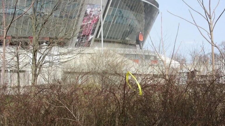 Ein gelbes Band hängt in den Zweigen eines Busches im blattlosen Buschwerk vor einem Stadion fest.