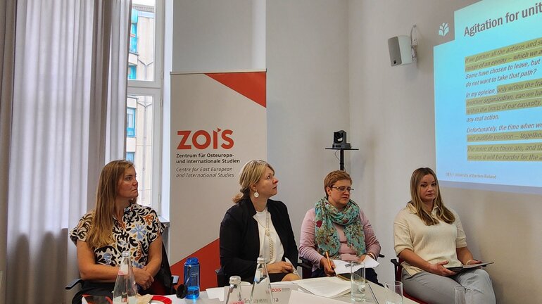 Four women are sitting on chairs. They are situated in front of a presentation projected at a wall and a poster with the ZOiS logo on it.