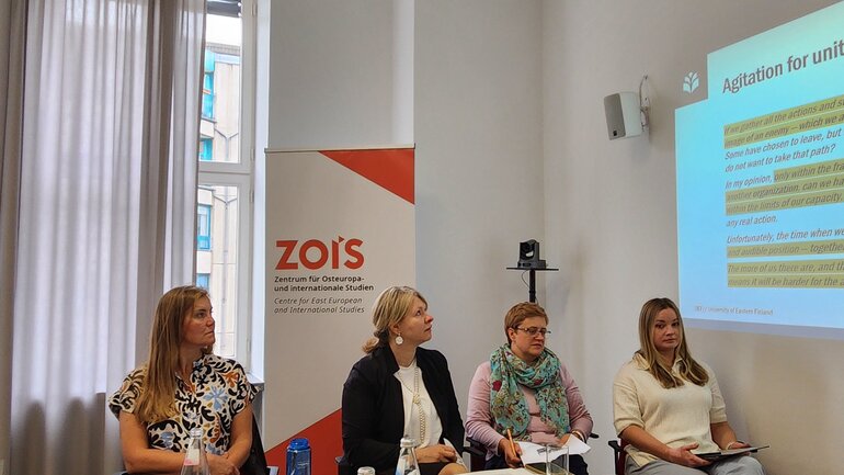Four women are sitting on chairs. They are situated in front of a presentation projected at a wall and a poster with the ZOiS logo on it.