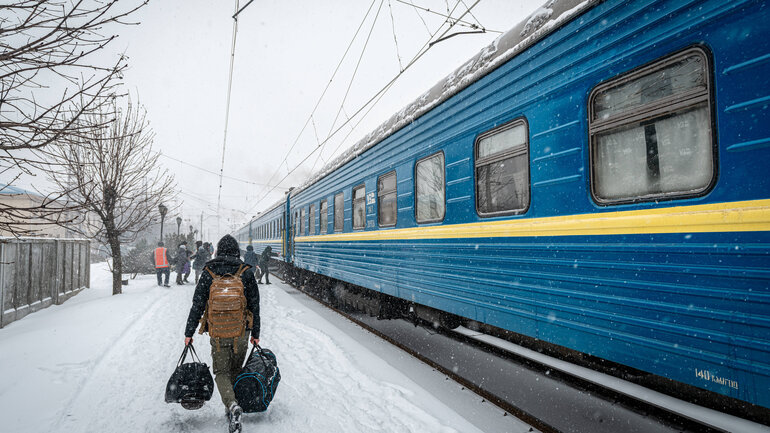 Ein Mann läuft mit Taschen bepackt an einem Bahnsteig in der Ukraine neben einem stehenden blau-weißen Zug.