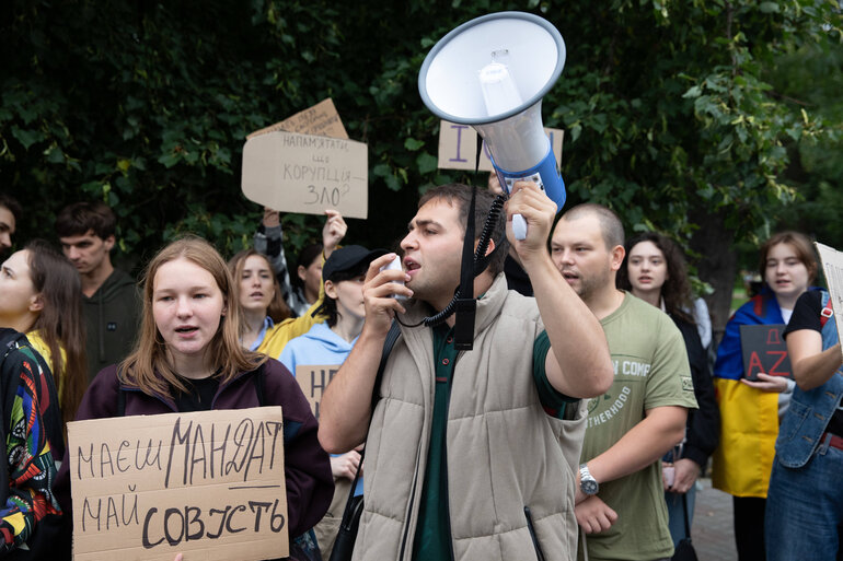 Eine Demonstration im Außenbereich. Im Vordergrund des Foto spricht eine Person in ein Megafon, neben ihr steht eine andere Person mit einem Protestschild, hinter ihnen sind weitere Demonstrierende.