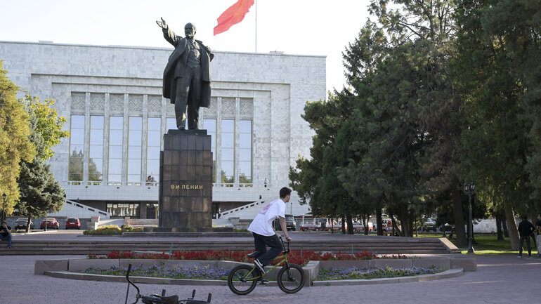 A child riding their bike in front of a Lenin monument, a red flag can be seen in the background on top of a large building.