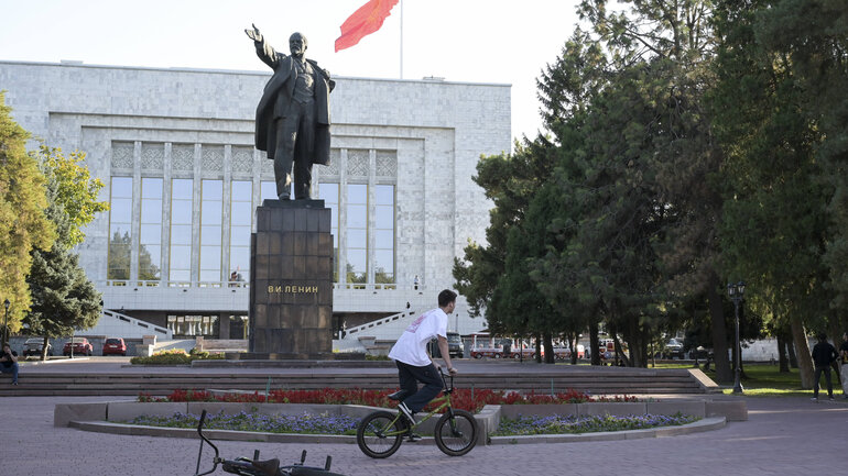 Ein Kind fährt mit seinem Fahrrad an einem Lenin Denkmal vorbei, im Hintergrund weht eine rote Fahne auf dem Dach eines großen Gebäudes.
