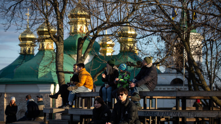 People are sitting on benches; in the background, a church with green and gold spires can be seen.