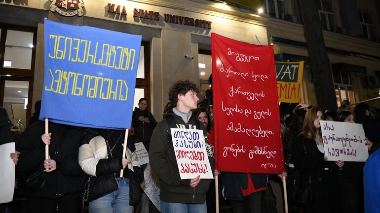 A group of young protesters is standing with signs in front of a building at Ilia State University.