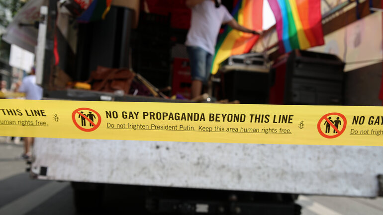Auf einem gelben Absperrband steht: „No gay propaganda beyond this line. Do not frighten President Putin. Keep this area free of human rights". Eine Regenbogenfahne mit Menschen auf einem Lastwagen ist im Hintergrund verschwommen zu sehen.