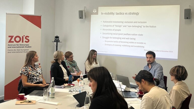 A group of people is situated sitting at a table in front of a presentation prjected at a wall.
