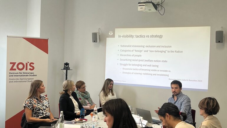 A group of people is situated sitting at a table in front of a presentation prjected at a wall.