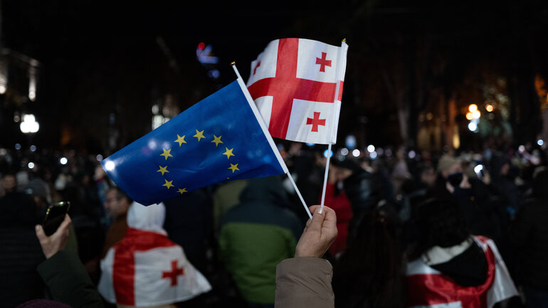 Eine Hand hält eine EU- und eine Georgien-Flagge. Im Hintergrund ist eine Menge Demonstrierender zu sehen.