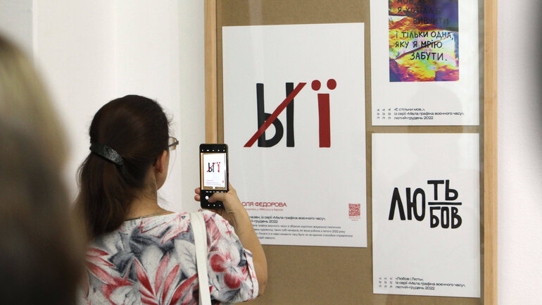 A Russian letter on a sign has been crossed out and replaced by a Ukrainian one. A woman takes a photo of it with a smartphone.