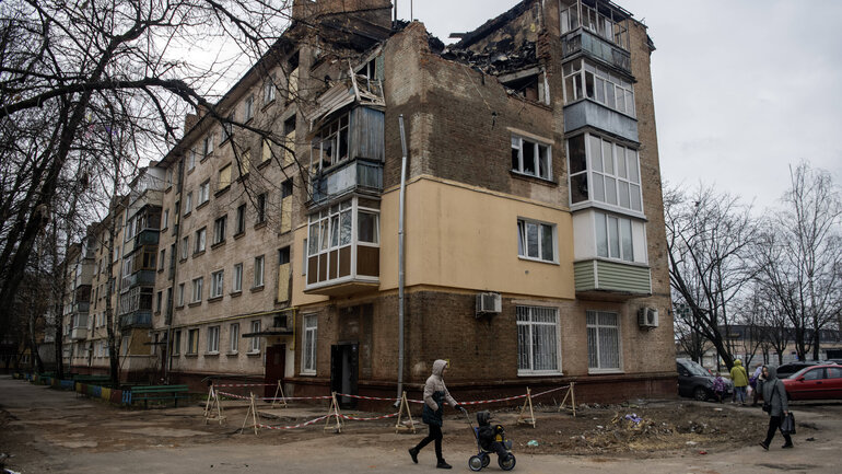A grey apartment building on the corner of a block of flats. The house is completely destroyed up to the second floor, a large part of the roof has been torn off.