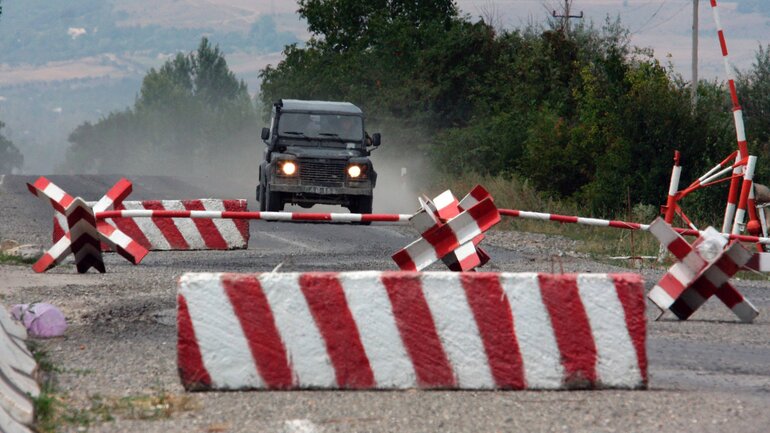 An off-road vehicle drives along a crumbling asphalt road towards red and white painted anit-tank barriers.