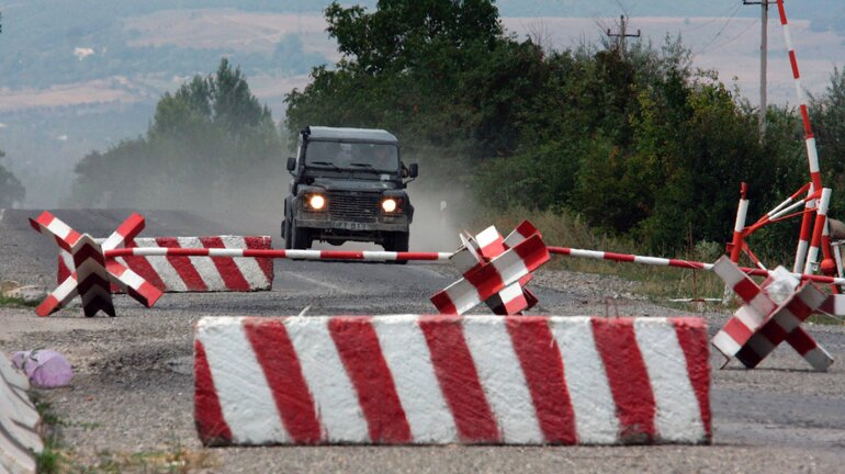 Ein Geländewagen fährt auf einer bröckelnden Asphaltstraße auf rot und weiß gestrichene Panzersperren zu.