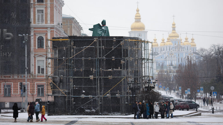 A statue towers above a protective wall built around it; in the background, a church with golden domes can be seen.