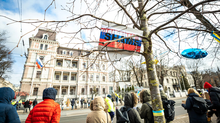 A group of people is gathered on the side of a road in front of a large building. Above them, a Russian flag reading "shame" is stuck in the branches of a tree.