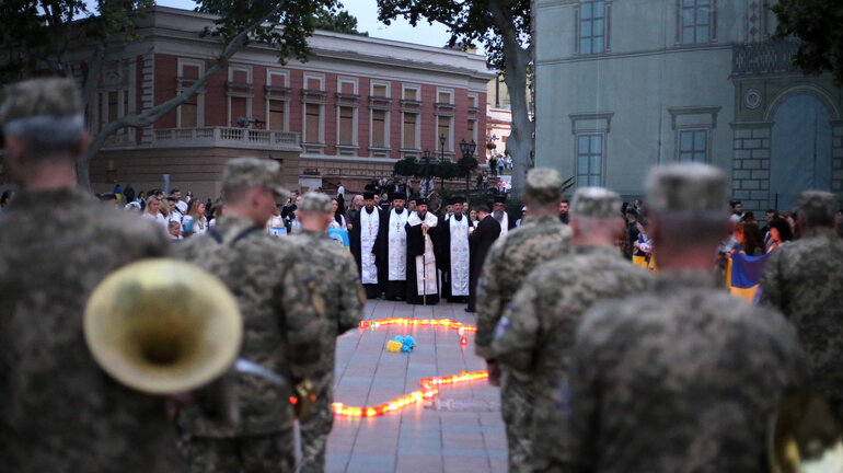 Mit dem Rücken zur Kamera stehen Männer in Militäruniformen, im Hintergrund sind orthodoxe Geistliche zu sehen, deren Blicke ebenfalls auf eine Zahl Kerzen und blau-gelbe Blumen gerichtet sind, welche zwischen den beiden Gruppen auf dem Boden liegen.