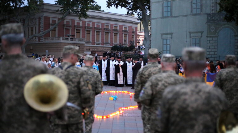 Men in military uniforms are standing with their backs to the camera, and in the background, Orthodox clergy can be seen, their gazes also directed at a number of candles and blue-and-yellow flowers lying on the ground between the two groups.