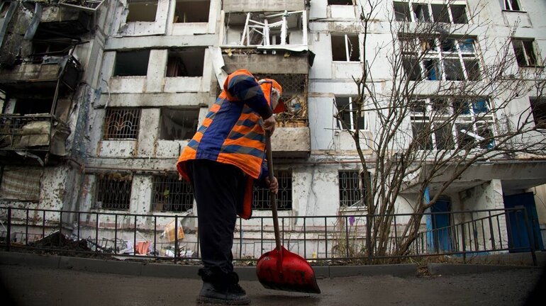 Damaged residential building after a drone attack, with street cleaners working among the rubble and debris in front of it.