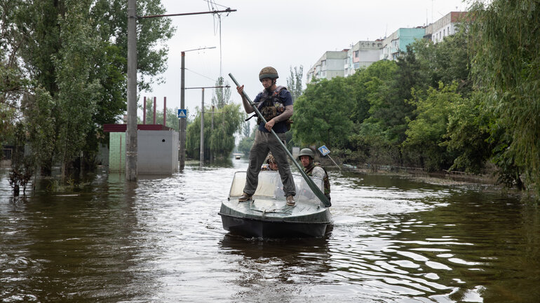 A man with an oar stands on a boat travelling through a flooded street.