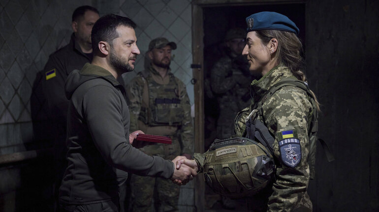 Ukrainian President Volodymyr Zelensky shakes hands with a female marine, with two soldiers standing in the background.