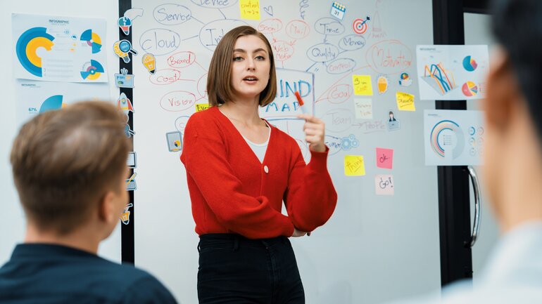 Eine junge Frau steht vor einem Smartboard und erklärt ihren Zuhörenden etwas.