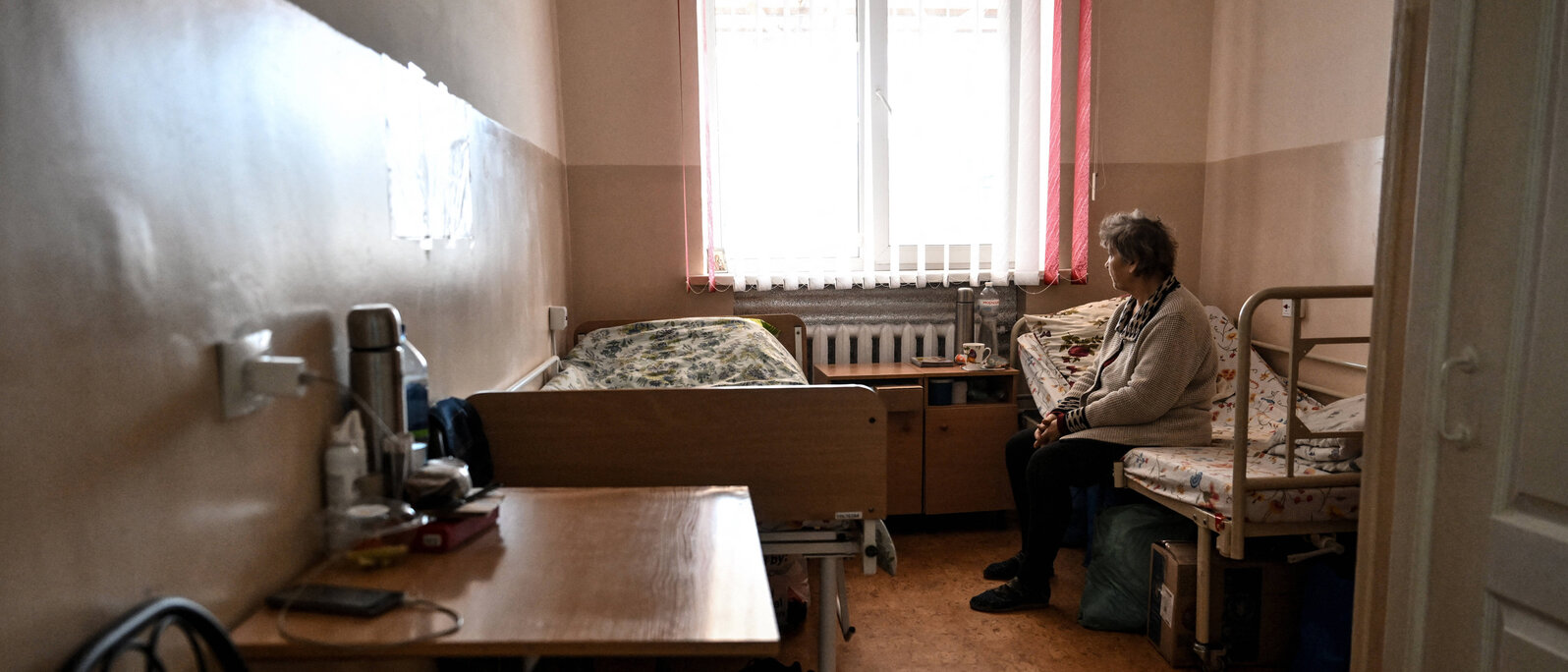 Eine ältere Frau sitzt in einem schlecht beleuchteten Zimmer auf einem von zwei Betten und sieht aus dem Fenster.