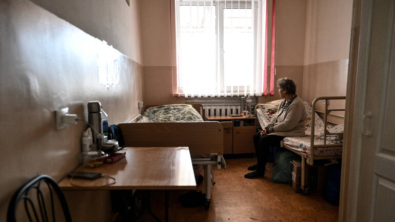Eine ältere Frau sitzt in einem schlecht beleuchteten Zimmer auf einem von zwei Betten und sieht aus dem Fenster.