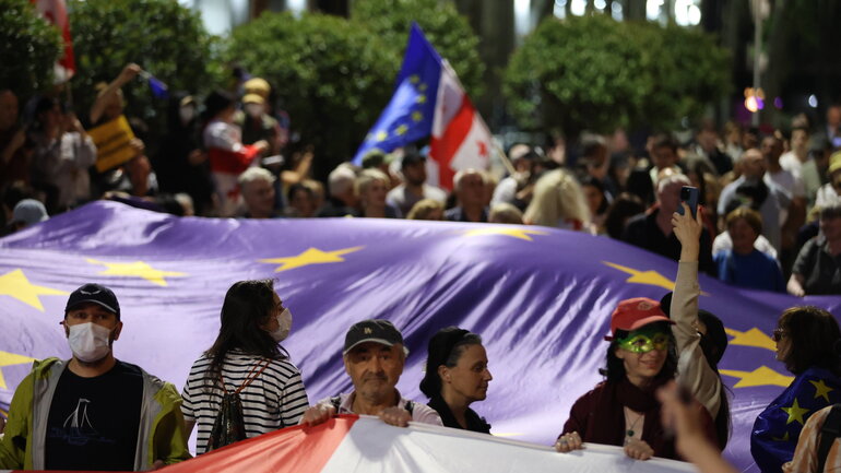 Photo of a protest outside. In the centre, behind a row of people, a very large EU flag is held horizontally above the ground. In the background, a large group of people are gathered.