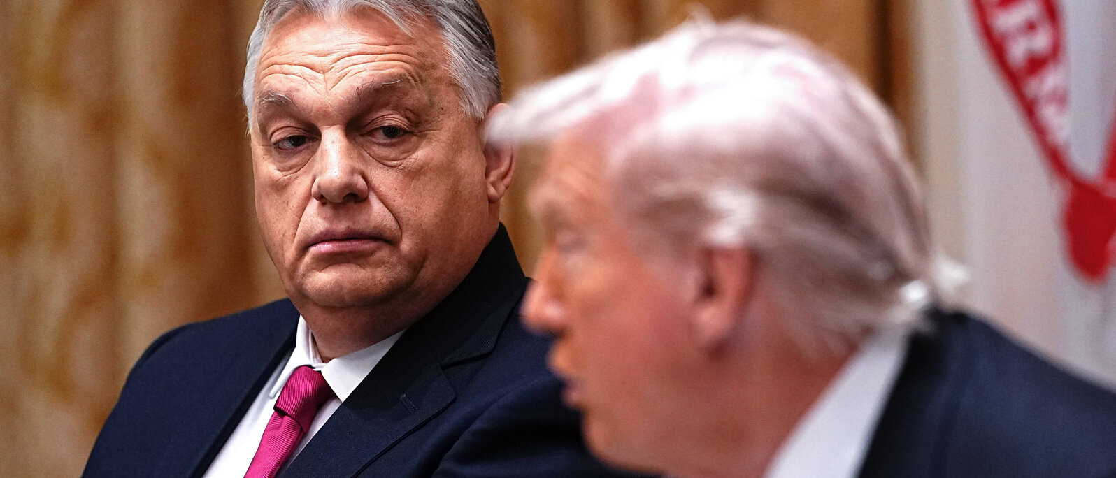 Viktor Orbán steht im Fokus des Bildes und sieht den grade sprechenden Donald Trump mit ernstem Blick an.