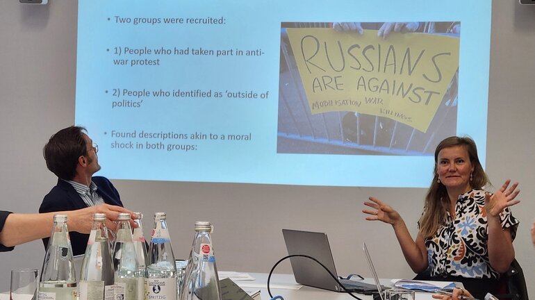 A woman and a man are sitting on chairs at a table in front of a presentation that is projected at a wall behind them.