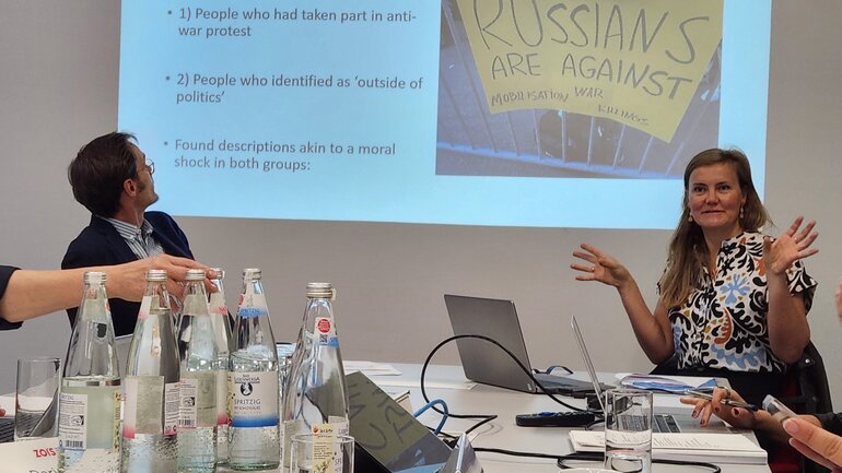 A woman and a man are sitting on chairs at a table in front of a presentation that is projected at a wall behind them.